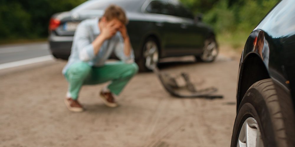 Man crouching on road after car accident, with damaged vehicle in background.
